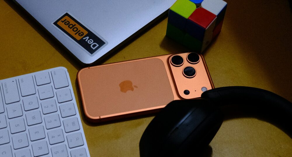 Laptop, phone, keyboard, headphones, and puzzle cube on desk.