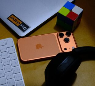 Laptop, phone, keyboard, headphones, and puzzle cube on desk.