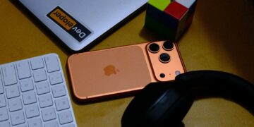 Laptop, phone, keyboard, headphones, and puzzle cube on desk.