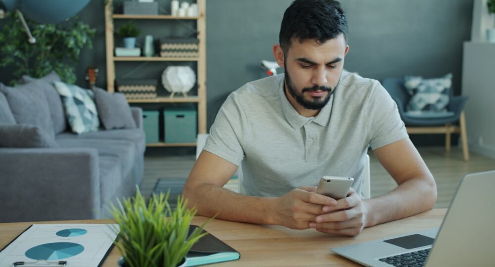 Man using smartphone at desk with laptop and charts.
