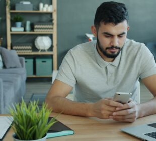Man using smartphone at desk with laptop and charts.