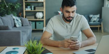 Man using smartphone at desk with laptop and charts.