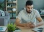 Man using smartphone at desk with laptop and charts.