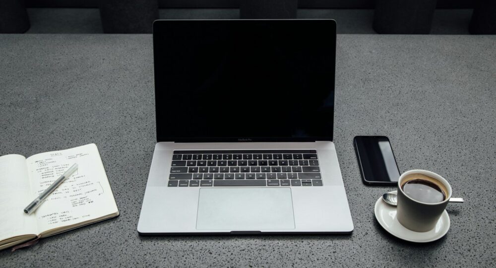 turned-off MacBook Pro between cup of coffee, iPhone, notebook, and pen